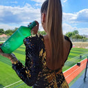 Vodka Voronskaya Bio - Person in a black and gold sequined dress holding a green bottle on a sports field.