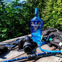 Vodka Voronskaya Blue - Blue bottle of Vodka Voronskaya with a camera and hiking equipment on a wooden surface with a forest background